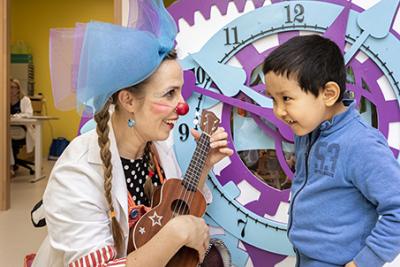 A hospital clown plays the guitar with a young patient. A hospital staff member sits in an office in the background.