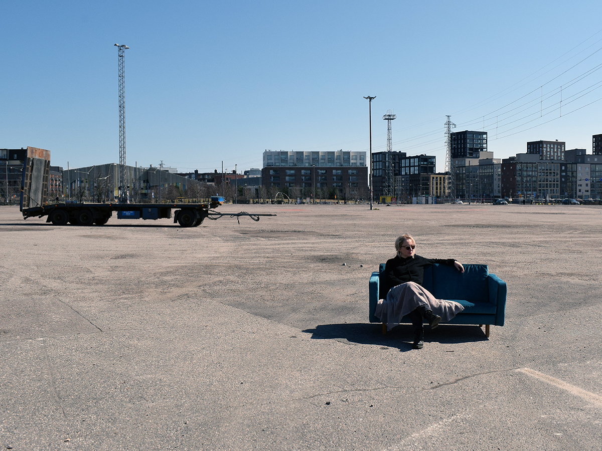 Pilvi Porkola is sitting on a blue sofa on tarmack. Behind her in the background there is a trailer chassis and apartment buildings.